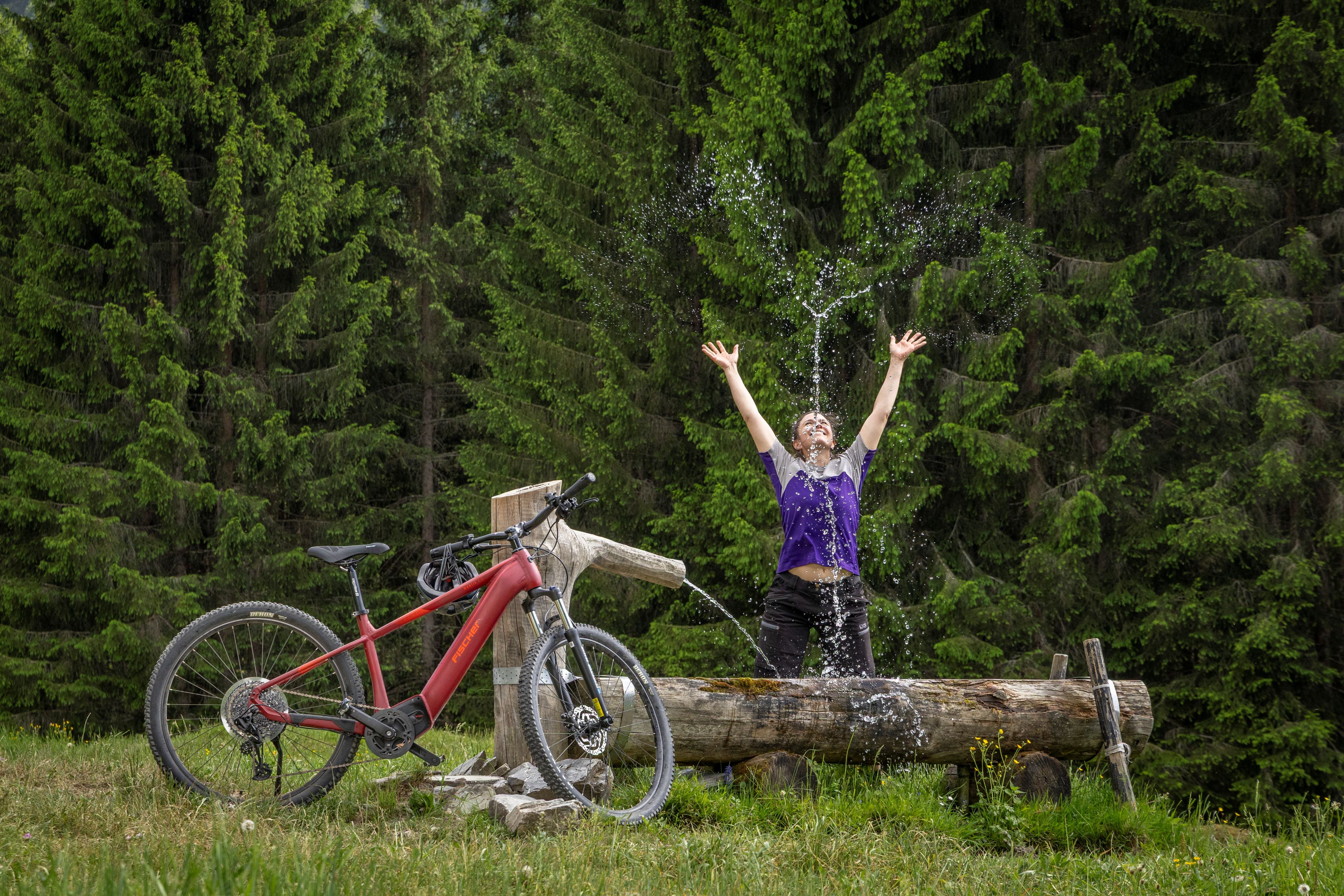 Eine Frau freut sich über einen Trinkbrunnen in den Bergen. Nach einer Mountainbike Tour mit ihrem E-Mountainbike MONTIS von Fischer. 